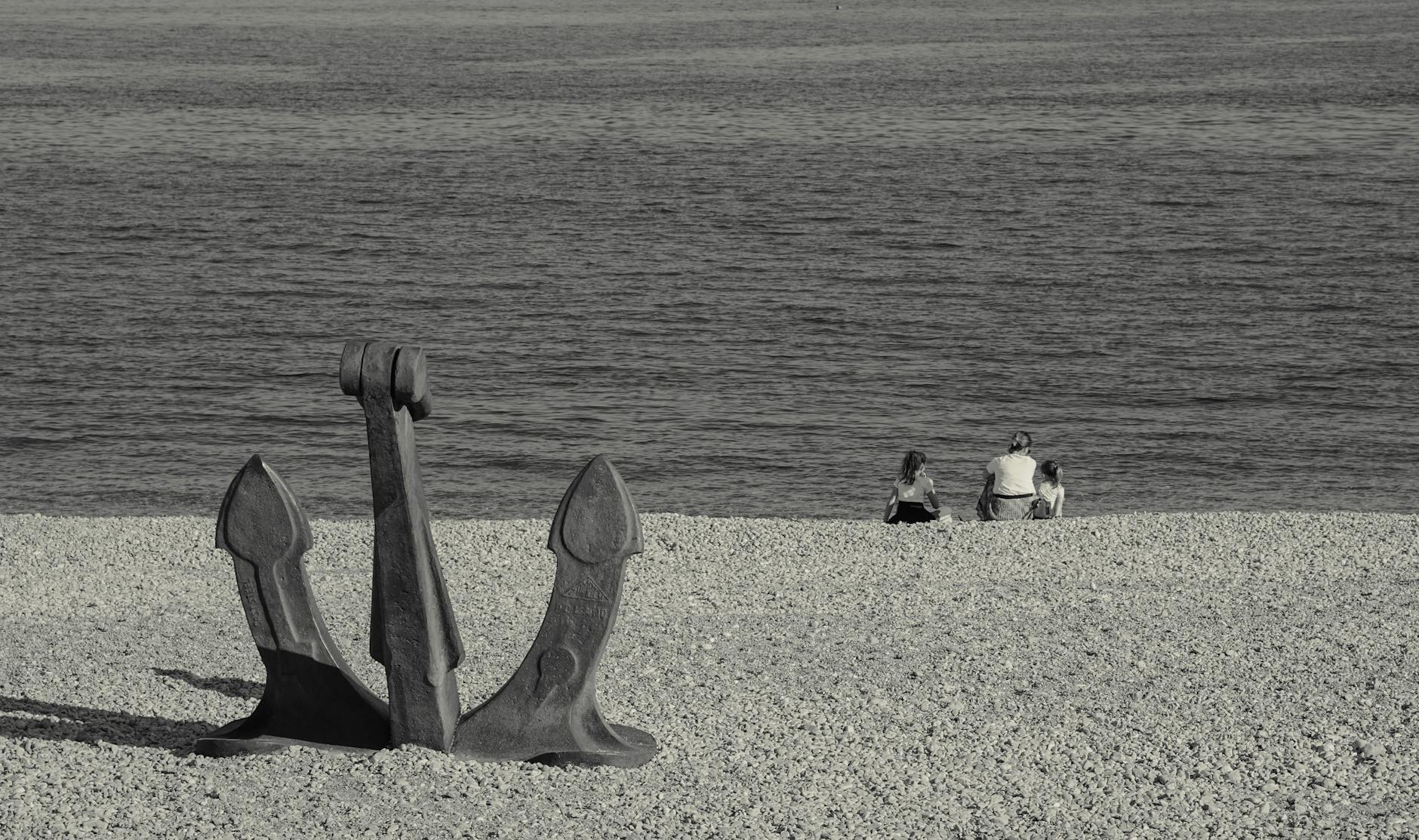 Rodzina z dziećmi siedzi przy morzu obok dużej kotwicy na kamienistej plaży